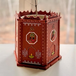 May include: A red, hexagonal planter with ornate white detailing and illustrations of deities. The planter holds a green plant with thin stems and leaves. The planter is placed in front of a window.