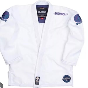 May include: White martial arts uniform with the text "CLASSIC" and "SHOYOROLL" in purple. The uniform has patches with circular designs in blue and purple. The gi is made of a durable material and is designed for training and competition.