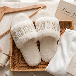 Könnte beinhalten: Ein Brautgeschenk-Set mit flauschigen weißen Hausschuhen mit silbernem "BRIDE"-Schriftzug, einem Holzkleiderbügel mit "MAID OF HONOR", einem weißen Bademantel und "Thank You"-Karten. Die Artikel sind in einem Weidenkorb angeordnet.