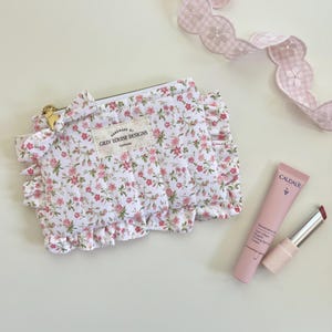 May include: A white floral print cosmetic bag with pink roses and ruffles, a gold zipper, and the text "HANDMADE BY GILLY LOUISE DESIGNS California". A pink eye cream tube and lipstick are next to the bag. A pink and white ribbon is in the background.