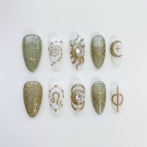 May include: A set of ten artificial nails with celestial designs. The nails feature gold accents on a variety of colors, including sage green and white. Designs include stars, suns, moons, and zodiac symbols. The nails are oval-shaped.