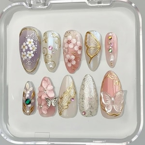 Könnte beinhalten: Ein Set aus zehn künstlichen Nägeln in einer transparenten Kunststoffbox. Die Nägel weisen verschiedene Designs auf, darunter Blumenmuster, Schmetterlinge und goldfarbene Akzente. Zu den Farben gehören Rosa, Weiß, Gold und Lila. Einige Nägel haben Strasssteine und Perlen.