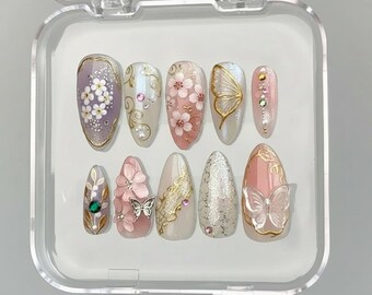 Op de afbeelding: Een set van tien kunstnagels in een doorzichtige plastic doos. De nagels hebben verschillende ontwerpen, waaronder bloempatronen, vlinders en gouden accenten. Kleuren zijn onder meer roze, wit, goud en paars. Sommige nagels hebben strass en parels.