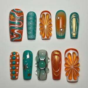 May include: Ten artificial nails with diverse designs. The nails showcase bright colours such as orange, teal, and gold, with patterns including swirls, dots, stars, and floral motifs. Some nails have a glossy finish, while others have a textured appearance.