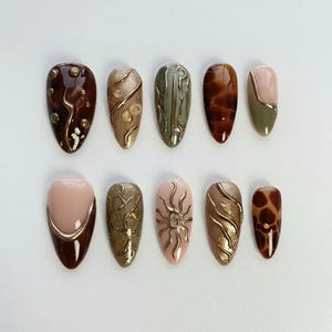 Op de afbeelding: Een verzameling van tien kunstnagels in verschillende ontwerpen. De nagels hebben een reeks kleuren, waaronder bruin, goud, groen en roze. Sommige nagels hebben textuurpatronen, terwijl andere gouden accenten of een zonontwerp hebben. De nagels zijn ovaal van vorm.
