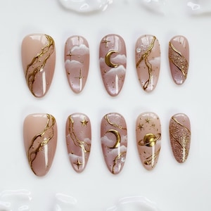 Puede incluir: Un conjunto de diez uñas postizas en forma de almendra. Las uñas están pintadas en un color rosa nude con detalles dorados. Los diseños incluyen espirales doradas, lunas, estrellas, nubes y un planeta. Las uñas están dispuestas sobre una superficie blanca.