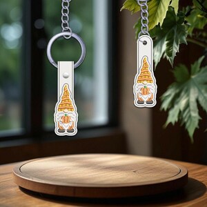 May include: Two keychains with a gnome design. The gnomes have white beards, orange hats, and are holding carrots. The keychains are attached to silver chains and white leather straps. The background includes a wooden table and green foliage.