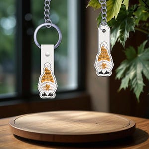 May include: Two keychains with a gnome design. The gnomes have orange and yellow striped hats, white beards, and are holding a leaf. The keychains are attached to a silver chain and a white leather strap.