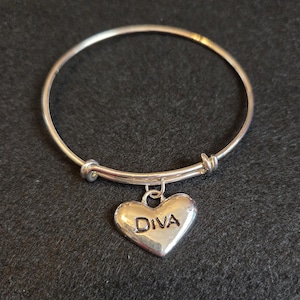 May include: Silver-toned bangle bracelet with a heart-shaped charm. The charm is engraved with the word "DIVA". The bracelet is displayed on a dark gray surface.