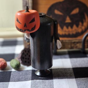 May include: A black wooden Halloween figure with a carved orange pumpkin head. The pumpkin has a jack-o'-lantern face. The figure is on a black and white checkered surface. A wooden sign with the word "Halloween" is in the background.