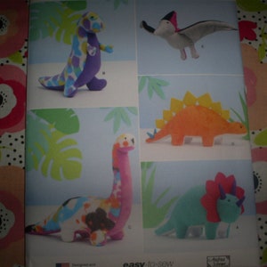 May include: A sewing pattern for five plush dinosaur toys. The pattern includes a purple and white dinosaur, a green and white dinosaur, a orange dinosaur with a yellow spine, a blue and red dinosaur, and a pink and white pterodactyl.