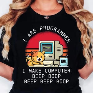 K&ouml;nnte beinhalten: Schwarzes T-Shirt mit einer Grafik, die einen Retro-Computer, eine l&auml;chelnde orangefarbene Katze und den Text "I ARE PROGRAMMER. I MAKE COMPUTER BEEP BOOP BEEP BEEP BOOP" in Wei&szlig; zeigt. Das Design beinhaltet einen roten und orangen Sonnenuntergang.