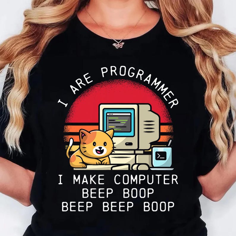 K&ouml;nnte beinhalten: Schwarzes T-Shirt mit einer Grafik, die einen Retro-Computer, eine l&auml;chelnde orangefarbene Katze und den Text "I ARE PROGRAMMER. I MAKE COMPUTER BEEP BOOP BEEP BEEP BOOP" in Wei&szlig; zeigt. Das Design beinhaltet einen roten und orangen Sonnenuntergang.
