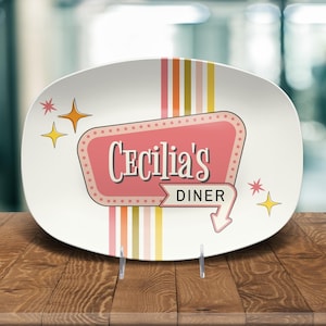 May include: An oval white serving platter featuring a pink retro-style sign with the text Cecilia's DINER in dark lettering. The design includes vertical stripes in muted tones of pink, orange, and yellow, accented by small yellow and pink star shapes.