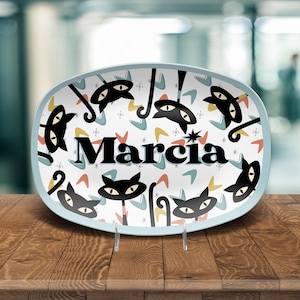 May include: An oval ceramic serving platter with a light blue border. The platter features a white background with black cat illustrations, starbursts, and colorful boomerang shapes. The name "Marcia" is printed in large black letters.