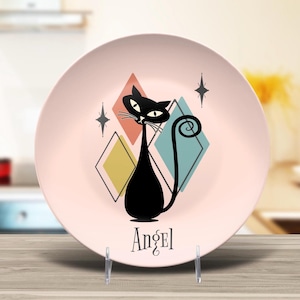 May include: A pink decorative plate featuring a black cat illustration with a curled tail, set against a backdrop of colorful diamond shapes. The word "Angel" is written below the cat in a stylized font.