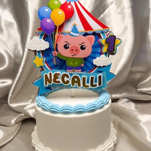 Puede incluir: Un pastel blanco con un adorno de tem&aacute;tica circense que presenta un cerdo de dibujos animados con un gorro de fiesta, sosteniendo globos de colores frente a una carpa a rayas rojas y blancas. Una pancarta azul muestra el nombre NECALLI en oro.