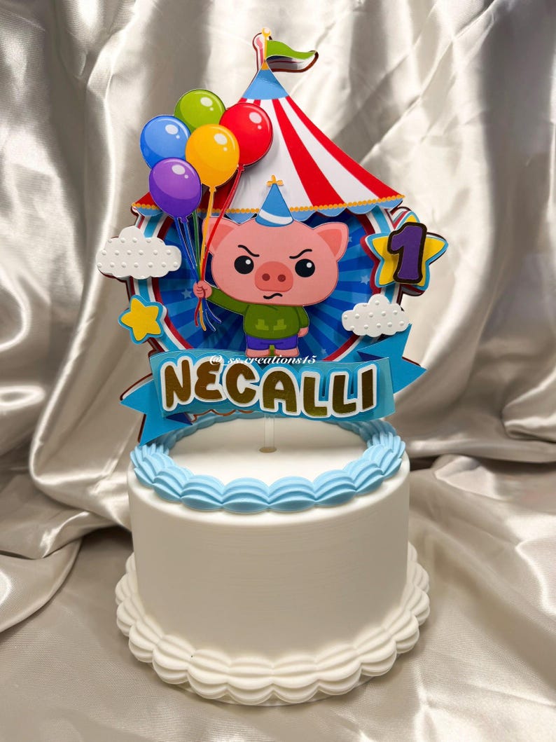 Puede incluir: Un pastel blanco con un adorno de tem&aacute;tica circense que presenta un cerdo de dibujos animados con un gorro de fiesta, sosteniendo globos de colores frente a una carpa a rayas rojas y blancas. Una pancarta azul muestra el nombre NECALLI en oro.