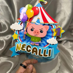 Puede incluir: Un adorno para pastel de varias capas que presenta un cerdo de dibujos animados con un sombrero de fiesta azul, sosteniendo un grupo de globos coloridos. El dise&ntilde;o incluye una carpa de circo a rayas rojas y blancas, nubes blancas, una estrella amarilla, un n&uacute;mero uno morado y una pancarta azul con el nombre NECALLI y el texto @ss.creations15.