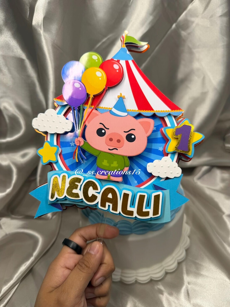 Puede incluir: Un adorno para pastel de varias capas que presenta un cerdo de dibujos animados con un sombrero de fiesta azul, sosteniendo un grupo de globos coloridos. El dise&ntilde;o incluye una carpa de circo a rayas rojas y blancas, nubes blancas, una estrella amarilla, un n&uacute;mero uno morado y una pancarta azul con el nombre NECALLI y el texto @ss.creations15.