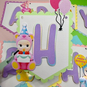 May include: A colorful birthday banner with the letter "H" in purple with gold trim. A pink and yellow bear-like figure holds a cake. Balloons in pink, blue, and purple are attached to the banner. The banner has a scalloped edge.
