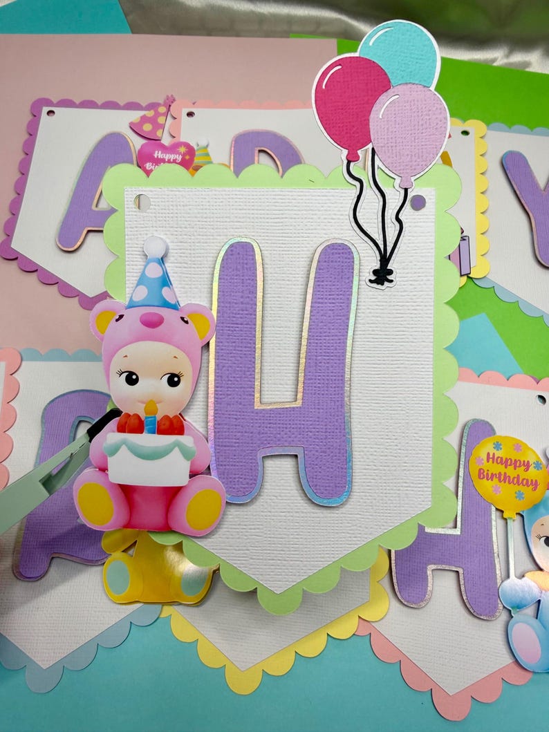 May include: A colorful birthday banner with the letter "H" in purple with gold trim. A pink and yellow bear-like figure holds a cake. Balloons in pink, blue, and purple are attached to the banner. The banner has a scalloped edge.