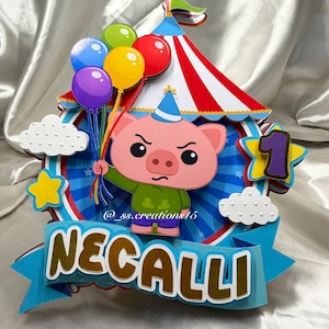 Puede incluir: Un adorno para tarta de papel multicapa que presenta un cerdo de dibujos animados con un sombrero de fiesta azul y una camisa verde, sosteniendo un racimo de globos de colores. El dise&ntilde;o incluye una carpa de circo a rayas rojas y blancas, nubes blancas, una estrella amarilla, un n&uacute;mero 1 morado y una pancarta azul con el nombre NECALLI en letras doradas.