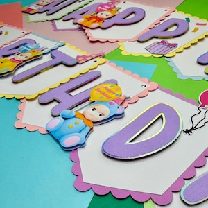 May include: A colorful birthday banner with the words "HAPPY BIRTHDAY" in purple letters. Each letter is on a white scalloped edge card. The banner includes cartoon baby figures and balloons. The background is a mix of pastel colors.