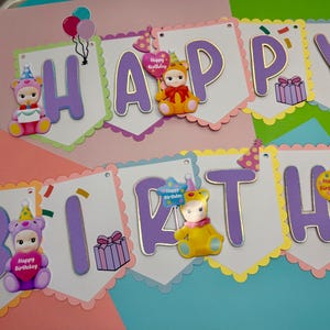 May include: A colorful birthday banner with the words "HAPPY BIRTHDAY" in purple letters. The banner is decorated with cute bear-like figures, balloons, and gift boxes. The background is a mix of pink, green, and blue.