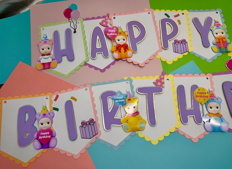 May include: A colorful birthday banner with the words "HAPPY BIRTHDAY" in purple letters. The banner is decorated with cute bear-like figures, balloons, and gift boxes. The background is a mix of pink, green, and blue.