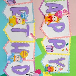 May include: A colorful birthday banner with the words "HAPPY BIRTHDAY" in purple letters. Each letter is on a white pennant with a scalloped edge, decorated with bear-shaped embellishments and confetti.