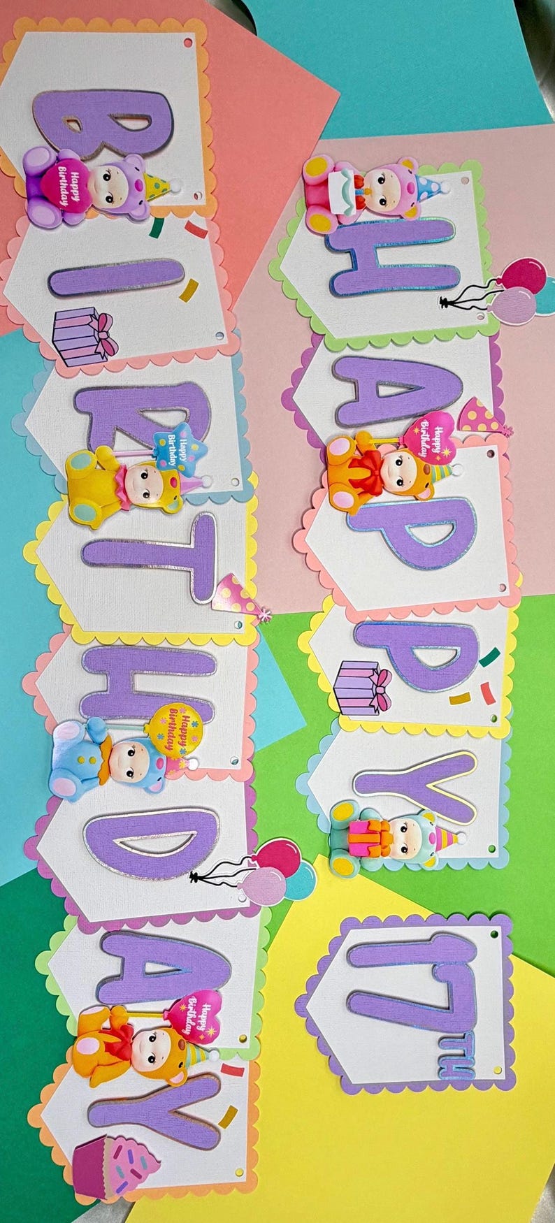 May include: A colorful birthday banner with the words "HAPPY BIRTHDAY" in purple letters. Each letter is on a white pennant with a scalloped edge, decorated with bear-shaped embellishments and confetti.