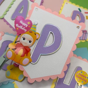 May include: A colorful birthday banner with scalloped edges and the letter "P" in purple. A cartoon bear holding a "Happy Birthday" balloon is attached. The banner is decorated with pastel colors, including pink, yellow, and blue.