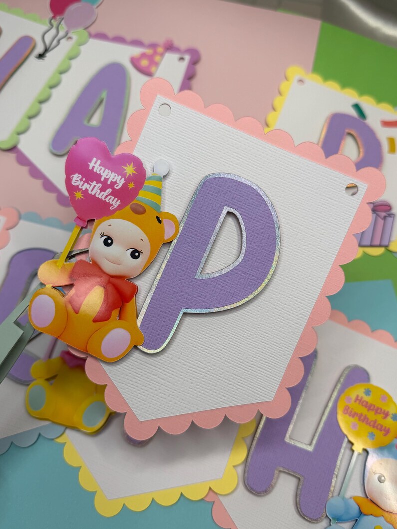 May include: A colorful birthday banner with scalloped edges and the letter "P" in purple. A cartoon bear holding a "Happy Birthday" balloon is attached. The banner is decorated with pastel colors, including pink, yellow, and blue.