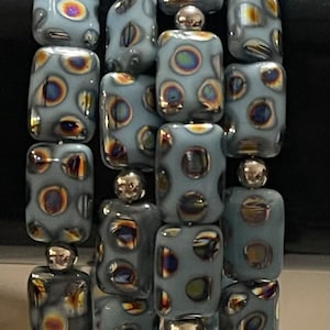 May include: A bracelet featuring multiple strands of light blue, rectangular beads. Each bead has a pattern of iridescent circles in shades of gold, brown, and green. The beads are separated by small, silver-colored beads.