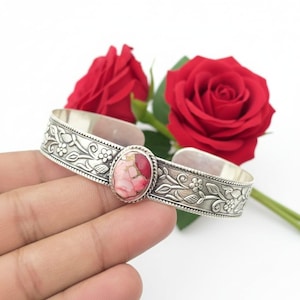May include: Silver cuff bracelet with an oval, pink and gold gemstone centre. The bracelet features a floral and vine design. Two red roses are in the background.
