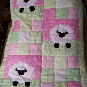 May include: A patchwork baby quilt featuring three white sheep appliqués with black faces and legs. The quilt is made of pink, green, and white patterned squares, with a green border. A charming and cozy nursery item.
