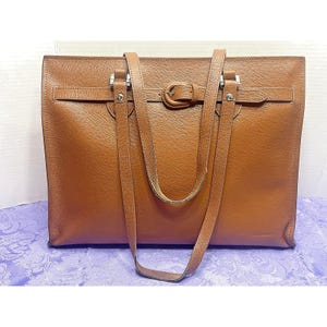May include: A tan leather tote bag featuring a horizontal strap across the front with a central buckle closure, silver-toned metal hardware, and two long shoulder straps attached with silver rivets. The bag has a rectangular shape and smooth finish.