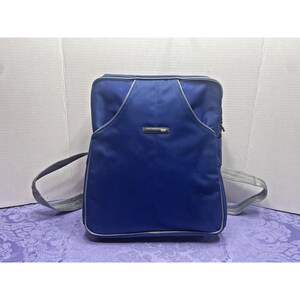 May include: A rectangular royal blue backpack featuring a textured fabric front panel with a central horizontal logo plaque. The bag includes light grey adjustable shoulder straps and silver piping accents along the edges of the main compartment.
