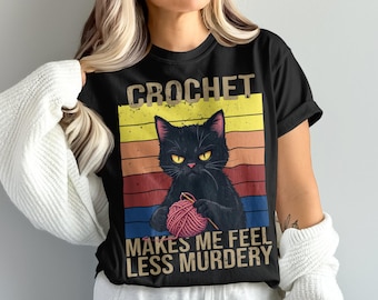 May include: Black t-shirt with a graphic of a black cat holding a ball of pink yarn. The text "CROCHET MAKES ME FEEL LESS MURDERY" is printed on the shirt in a distressed font, with a retro sunset background.