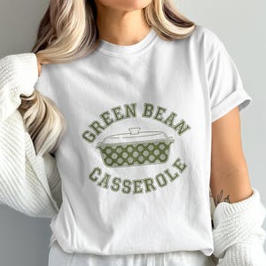 May include: White t-shirt featuring a green bean casserole design. The text "GREEN BEAN CASSEROLE" arches above and below a casserole dish illustration. The dish is green with a floral pattern.
