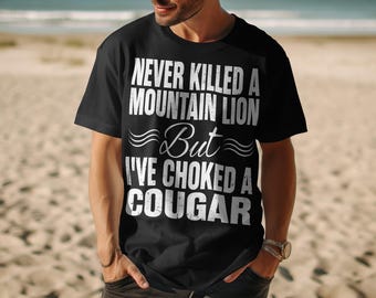 May include: Black t-shirt with white text that reads "NEVER KILLED A MOUNTAIN LION But I'VE CHOKED A COUGAR". The shirt is being worn by a person on a beach.