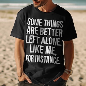 Puede incluir: Camiseta negra con el texto blanco: "ALGUNAS COSAS ES MEJOR DEJARLAS SOLAS. COMO YO, POR EJEMPLO." La camiseta es de cuello redondo y la lleva una persona. El fondo es una playa de arena.