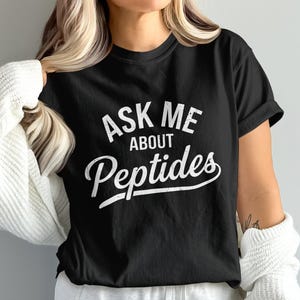 May include: A black short-sleeved t-shirt featuring the white distressed-style text "ASK ME ABOUT Peptides" printed on the front. The shirt has a crew neckline and a standard fit, with the word "Peptides" written in a larger, cursive font.