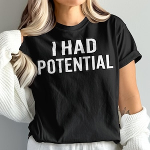 May include: Black t-shirt with the white text "I HAD POTENTIAL". The shirt is a crew neck style. The person is wearing a white sweater over the shirt.