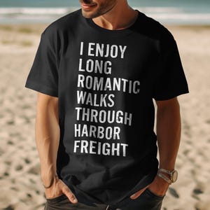 May include: Black t-shirt with white text that reads "I Enjoy Long Romantic Walks Through Harbor Freight." The shirt is a crew neck and is being worn by a person on a beach.