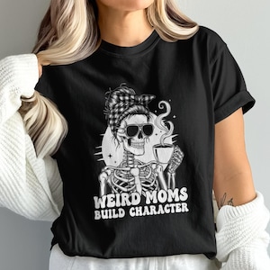May include: Black t-shirt with a skeleton wearing sunglasses and a bandana, holding a coffee cup. The text "WEIRD MOMS BUILD CHARACTER" is printed in white. The design is a humorous take on motherhood.