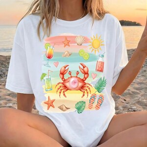 Op de afbeelding: Wit T-shirt met een strandthema. Het ontwerp bevat een rode krab met een kauwgomballenbel, cocktails, schelpen, een zon en slippers. Het kunstwerk is in aquarel stijl.