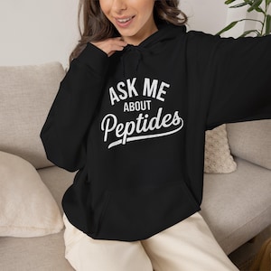 May include: A black long-sleeve hooded sweatshirt featuring a front kangaroo pocket and a drawstring hood. The chest displays white text that reads ASK ME ABOUT Peptides in a combination of block and script fonts with a distressed texture.