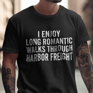 以下が含まれることがあります： 「I ENJOY LONG ROMANTIC WALKS THROUGH HARBOR FREIGHT」の白文字がプリントされた黒いTシャツ。クルーネックのTシャツです。Tシャツを着ている人は腕にタトゥーを入れています。